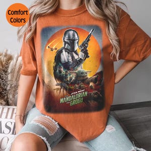 Op de afbeelding: Oranje T-shirt met een afbeelding van "The Mandalorian" met de Mandalorian met een blaster, Grogu en andere personages. De tekst "STAR WARS THE MANDALORIAN AND GROGU" staat eronder. Het shirt heeft de woorden "Comfort Colors" in een cirkel.