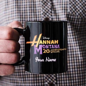 Puede incluir: Taza de cer&aacute;mica negra con el logotipo de "Hannah Montana" en dorado, morado y blanco. La taza incluye el texto "20th Anniversary Special" y espacio para un nombre personalizado. Una taza de caf&eacute; cl&aacute;sica.