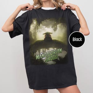 May include: Black t-shirt featuring a graphic from The Mandalorian and Grogu. The design shows Grogu in a forest setting. The Star Wars logo is in green and white. The shirt is a crew neck.