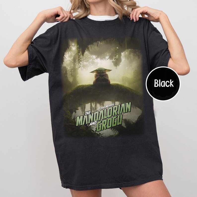 May include: Black t-shirt featuring a graphic from The Mandalorian and Grogu. The design shows Grogu in a forest setting. The Star Wars logo is in green and white. The shirt is a crew neck.