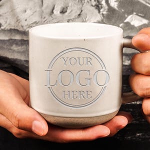 May include: A speckled off-white ceramic mug featuring a circular embossed design on the front with the text YOUR LOGO HERE centered inside. The mug has a smooth cylindrical body, a sturdy handle on the right side, and a slightly textured base.