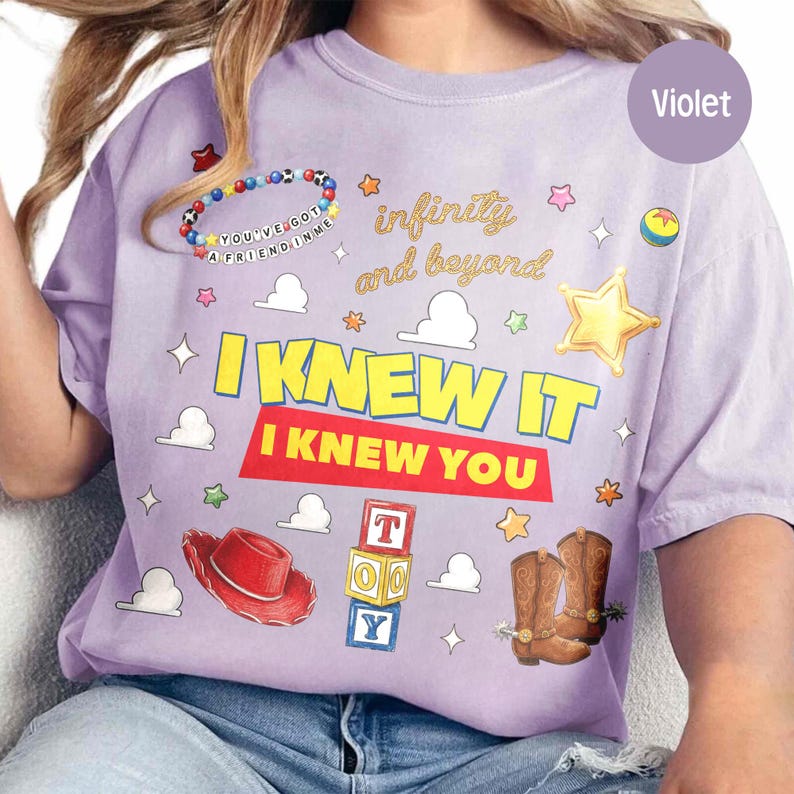 May include: A violet t-shirt featuring a graphic print with the text "I KNEW IT" and "I KNEW YOU" in yellow and red. The design includes a beaded bracelet, a red cowboy hat, brown cowboy boots, alphabet blocks, and cloud shapes.