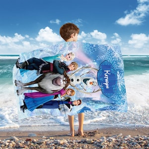 May include: A blue and white blanket featuring characters from the movie Frozen, including Elsa, Anna, Kristoff, Olaf, and Sven. The blanket has a snowflake design and the name "Kathryn" is printed on it. The blanket is being held up by a person on a beach.