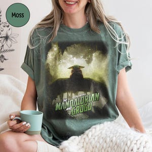 May include: Moss green t-shirt featuring a graphic from "The Mandalorian" with Grogu. The Star Wars logo is in green and white. The shirt is a relaxed fit and has rolled-up sleeves. A light blue mug is held in the hand.