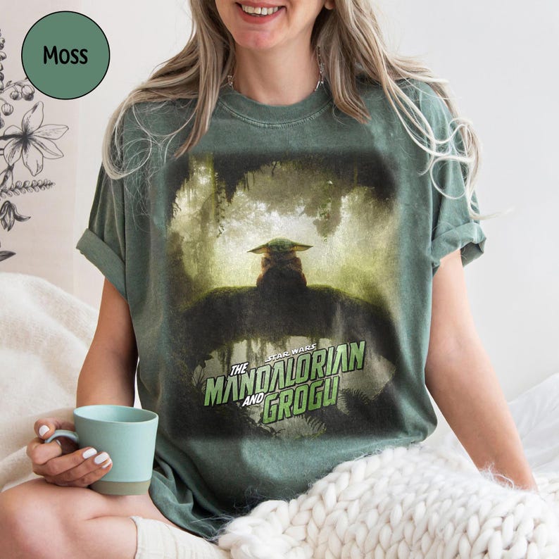 May include: Moss green t-shirt featuring a graphic from "The Mandalorian" with Grogu. The Star Wars logo is in green and white. The shirt is a relaxed fit and has rolled-up sleeves. A light blue mug is held in the hand.