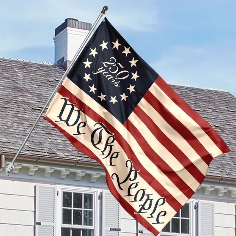 May include: A flag with a design reminiscent of the American flag, featuring red and white stripes and a blue field with stars. The flag displays the text "We The People" and "250 years". The flag is mounted on a pole and is displayed outside a house.