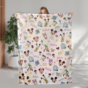 May include: A white blanket featuring a colorful pattern of Disney characters, including Mickey and Minnie Mouse, Donald Duck, and Goofy. The blanket also has the name "Sophia" written in a script font, along with various other Disney-themed icons.