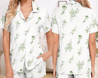 May include: White pajama set with a green botanical print. The set includes a short-sleeved button-down shirt and matching shorts. The shirt has black trim around the collar and buttons. The botanical pattern features green leaves and stems.