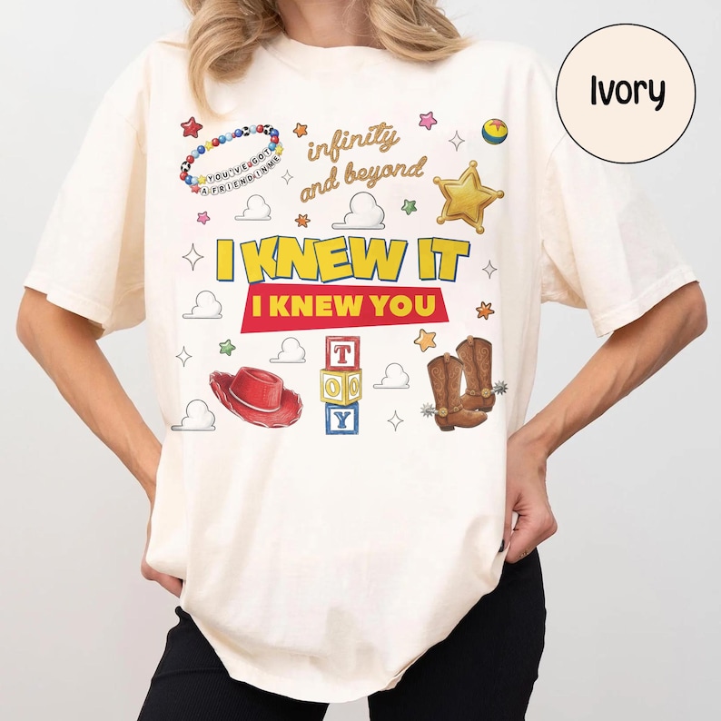 May include: An ivory t-shirt featuring a graphic print with the text "infinity and beyond", "I KNEW IT", and "I KNEW YOU". The design includes illustrations of a beaded bracelet, a gold star, a red cowboy hat, alphabet blocks, and cowboy boots.