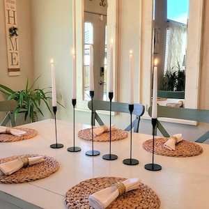 May include: A dining table set with six black metal candle holders, each holding a lit white candle. Woven placemats and folded napkins with gold napkin rings are arranged on the table. The table is white, and the room has a neutral colour palette.