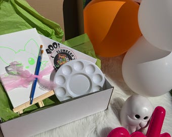 May include: A white box filled with Halloween-themed party supplies, including a white canvas with a ghost design, a paint palette, paintbrushes, a small easel, and a pink and white tulle ribbon. The box is surrounded by white, orange, and black balloons, and a pink "eek" sign.