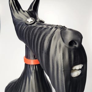 May include: A black and gray sculpture of a Scottish Terrier dog with an orange collar. The dog has white teeth and a button on its head. The sculpture is made of a ridged material.