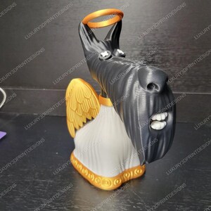 May include: A 3D-printed sculpture of a dog with angel wings and a halo. The dog is black with white teeth and a white chest. The wings and halo are gold, and the base is white with gold trim. The sculpture is a unique decorative item.