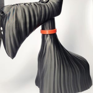 May include: A black Scottish Terrier figurine with an orange collar. The dog has a textured, ridged appearance and a small white button detail on its head. The figurine is set against a white background.
