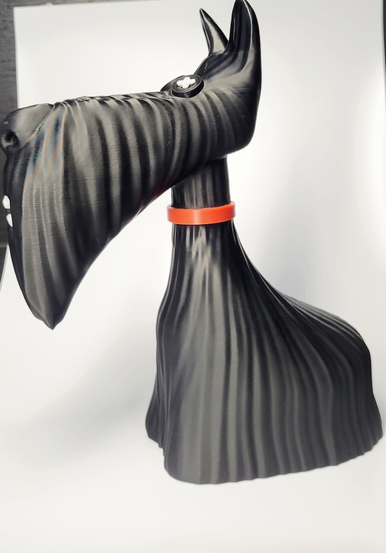 May include: A black Scottish Terrier figurine with an orange collar. The dog has a textured, ridged appearance and a small white button detail on its head. The figurine is set against a white background.