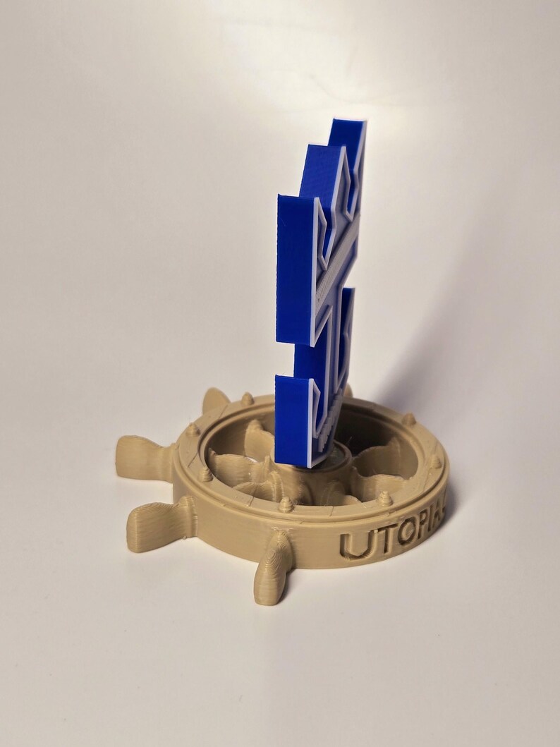 Peut inclure: Un logo bleu imprim&eacute; en 3D d'un gouvernail de navire avec le mot "Utopia" sur la base. Le logo est mont&eacute; sur un gouvernail de navire imprim&eacute; en 3D de couleur beige.