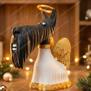 3D printed  Scottie dog figurine from Coraline, with a long black snout, white robe, golden wings, and a gold halo, displayed on a light wooden surface with bookshelves in the background. Measurement labels show 9.5 inches tall and 4 inches wide.”