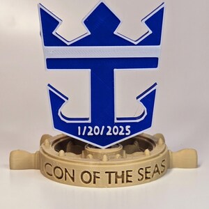 Peut inclure: Un logo de bateau de croisi&egrave;re Royal Caribbean imprim&eacute; en 3D bleu et blanc avec une couronne et une ancre. Le logo est mont&eacute; sur une base en 3D imprim&eacute;e marron avec les mots "CON OF THE SEAS" et la date "1/20/2025".