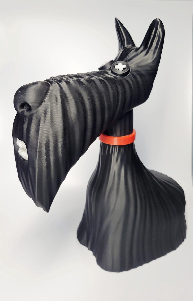 May include: A black Scottish Terrier figurine with a textured surface. The dog has a white mouth and a red collar. The figurine has a button-like detail on its head and is set against a white background.
