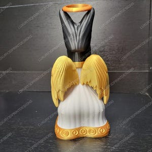 May include: A 3D-printed angel figurine with a black head and a gold halo. The angel has gold wings and a white robe with gold trim. The figurine is displayed on a dark surface.