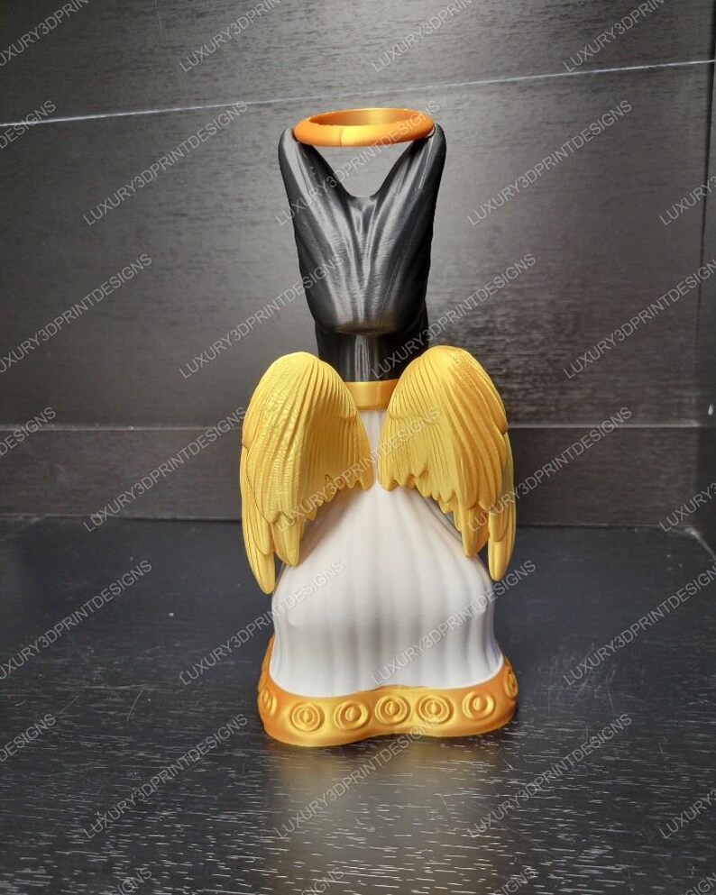 May include: A 3D-printed angel figurine with a black head and a gold halo. The angel has gold wings and a white robe with gold trim. The figurine is displayed on a dark surface.
