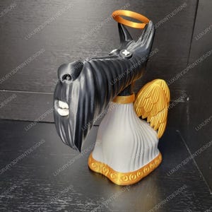 3D printed  Scottie dog figurine from Coraline, with a long black snout, white robe, golden wings, and a gold halo, displayed on a light wooden surface with bookshelves in the background. Measurement labels show 9.5 inches tall and 4 inches wide.”