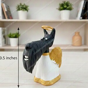 3D printed  Scottie dog figurine from Coraline, with a long black snout, white robe, golden wings, and a gold halo, displayed on a light wooden surface with bookshelves in the background. Measurement labels show 9.5 inches tall and 4 inches wide.”