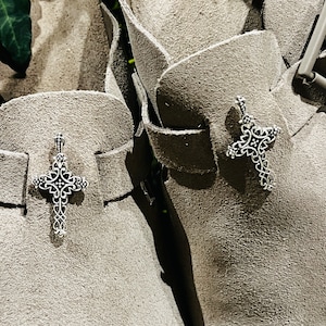 Könnte beinhalten: Zwei Wildlederstiefel mit silbernen Kreuzanhängern an den Riemen. Die Stiefel sind hellbraun und haben eine strukturierte Oberfläche.