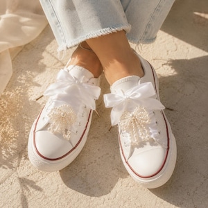 Op de afbeelding: Witte canvas sneakers met rode rand en parelversieringen. Elke schoen heeft een grote witte satijnen strik over de veters. De sneakers zijn een klassieke stijl, geschikt voor casual wear of speciale gelegenheden.