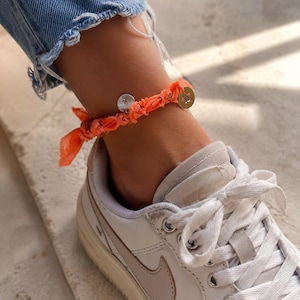 Op de afbeelding: Een oranje stoffen enkelbandje met twee kleine, ronde, zilverkleurige bedels en een goudkleurige sterbedel. De armband wordt gedragen boven een witte sneaker met een lichtbruine swoosh en witte veters. De persoon draagt een blauwe jeans.