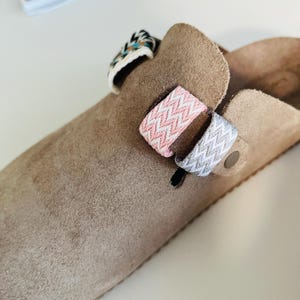 Può includere: Un paio di zoccoli in camoscio marrone con tre cinturini. I cinturini sono decorati con tessuto a chevron rosa e bianco e tessuto a motivo multicolore.