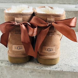 May include: A pair of brown suede UGG boots with a brown satin ribbon tied in a bow around the ankles. The boots have a white fur lining and the UGG logo is visible on the back of each boot.