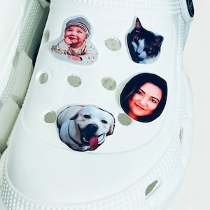 Op de afbeelding: Witte klompen versierd met vier kleurrijke, ronde stickers. De stickers tonen een lachende baby, een zwart-witte kat, een lachende vrouw en een gele Labrador Retriever met zijn tong uit.