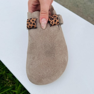 May include: A pair of light brown suede clogs with leopard print straps. The straps are adjustable and have a buckle closure.