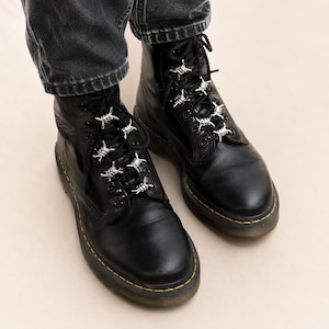 Könnte beinhalten: Schwarze Lederstiefel mit silbernen Stacheldraht-Schnürsenkel-Anhängern. Die Stiefel haben gelbe Nähte um die Sohlen und sind geschnürt. Getragen mit schwarzen Jeans.