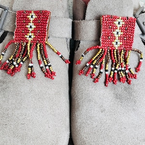 May include: A pair of gray suede shoes with red, gold, and black beaded tassels attached to the straps.