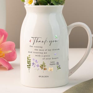 Pode incluir: Um vaso de cerâmica branco com uma alça, cheio de vegetação e uma flor de pompom amarela. O vaso tem uma mensagem impressa e detalhes florais. Outros vasos e flores estão no fundo.