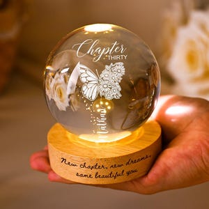 May include: A glowing glass sphere with a butterfly and floral design, resting on a wooden base. The sphere has the words "Chapter Thirty" and "Nathan" etched on it. The base reads "New chapter, new dreams, same beautiful you."