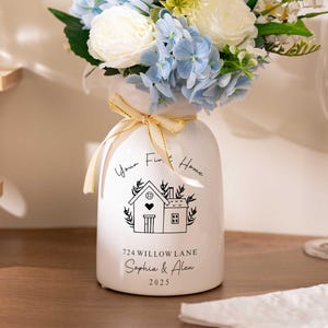 May include: A white ceramic vase with a floral arrangement of blue hydrangeas, white roses, and greenery. The vase has a house illustration and text that reads "Your First Home" and "724 Willow Lane, Sophia & Alex, 2025". A gold ribbon is tied around the vase.