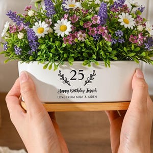 May include: A white rectangular ceramic planter with a bamboo base, filled with artificial purple lavender, white daisies, and small pink flowers. The planter features black text reading 25, Happy Birthday Joseph, and LOVE FROM MILA & AIDEN.