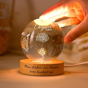 May include: A lit glass sphere with a floral design and the text "Chapter Thirty" and "Scarlett." The sphere sits on a wooden base with the inscription "New chapter, new dreams, same beautiful you."