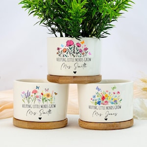 Puede incluir: Tres macetas de cerámica blanca con diseños florales y el texto "HELPING LITTLE MINDS GROW" y un nombre. Las macetas se asientan sobre platillos redondos de madera. Una maceta contiene una planta verde. Las macetas tienen ilustraciones coloridas de flores y mariposas.