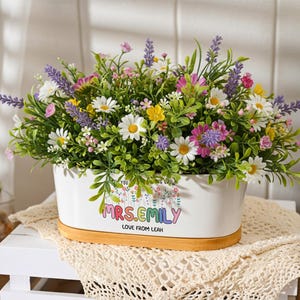 Puede incluir: Una jardinera de cerámica blanca llena de flores artificiales, incluyendo margaritas y lavanda. La jardinera tiene el texto "MRS. EMILY LOVE FROM LEAH" y se apoya en una base de madera. La composición floral es colorida.
