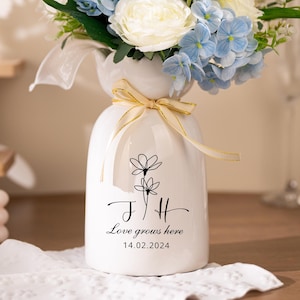 May include: A white ceramic vase with a bouquet of blue and white flowers, tied with a gold ribbon. The vase has a floral design and the text "Love grows here" with a date.