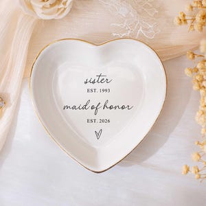 Op de afbeelding: Wit hartvormig sieradenschaaltje met gouden rand. Het schaaltje heeft de woorden "sister" en "maid of honor" erop gedrukt, samen met de jaartallen "EST. 1993" en "EST. 2026". Er is ook een klein hartsymbool aanwezig.