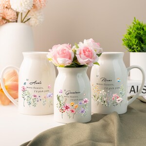 Op de afbeelding: Drie crèmekleurige keramische vazen en mokken met bloemstukken. De vazen en mokken hebben de zinnen "Als tantes bloemen waren, zou ik je kiezen", "Als oma's bloemen waren, zou ik je kiezen" en "Als mama's bloemen waren, zou ik je kiezen".