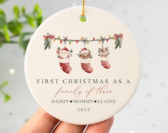 Può includere: Ornamento natalizio rotondo in ceramica con un design festivo. Presenta tre calze appese con Babbo Natale, un orso e una renna. Il testo recita "First Christmas as a family of three" e "2024". Include un cordino dorato per appenderlo.
