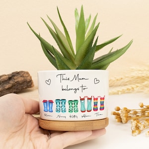 May include: White ceramic planter with the text "This Mum belongs to" and a heart on each side.  The planter has a row of colourful rain boots with names written below each boot:  "Kian", "Nancy", "Kathy", "Alan", and "Tina".  The planter sits on a wooden base.