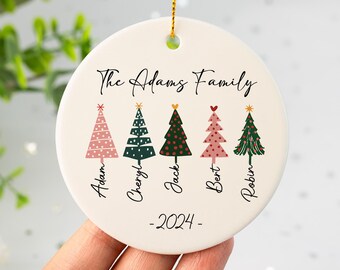 May include: A round, off-white ceramic Christmas ornament with a gold string. The ornament features the text "The Adams Family" above six stylized Christmas trees in pink and green, with names below. The year "-2024-" is printed at the bottom.