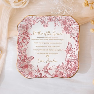 May include: A square, white ceramic trinket dish with a gold rim and floral design in pink. The dish has text that reads "Mother of the Groom" and a personal message. A gold ring with a large, light-colored stone is in the image.